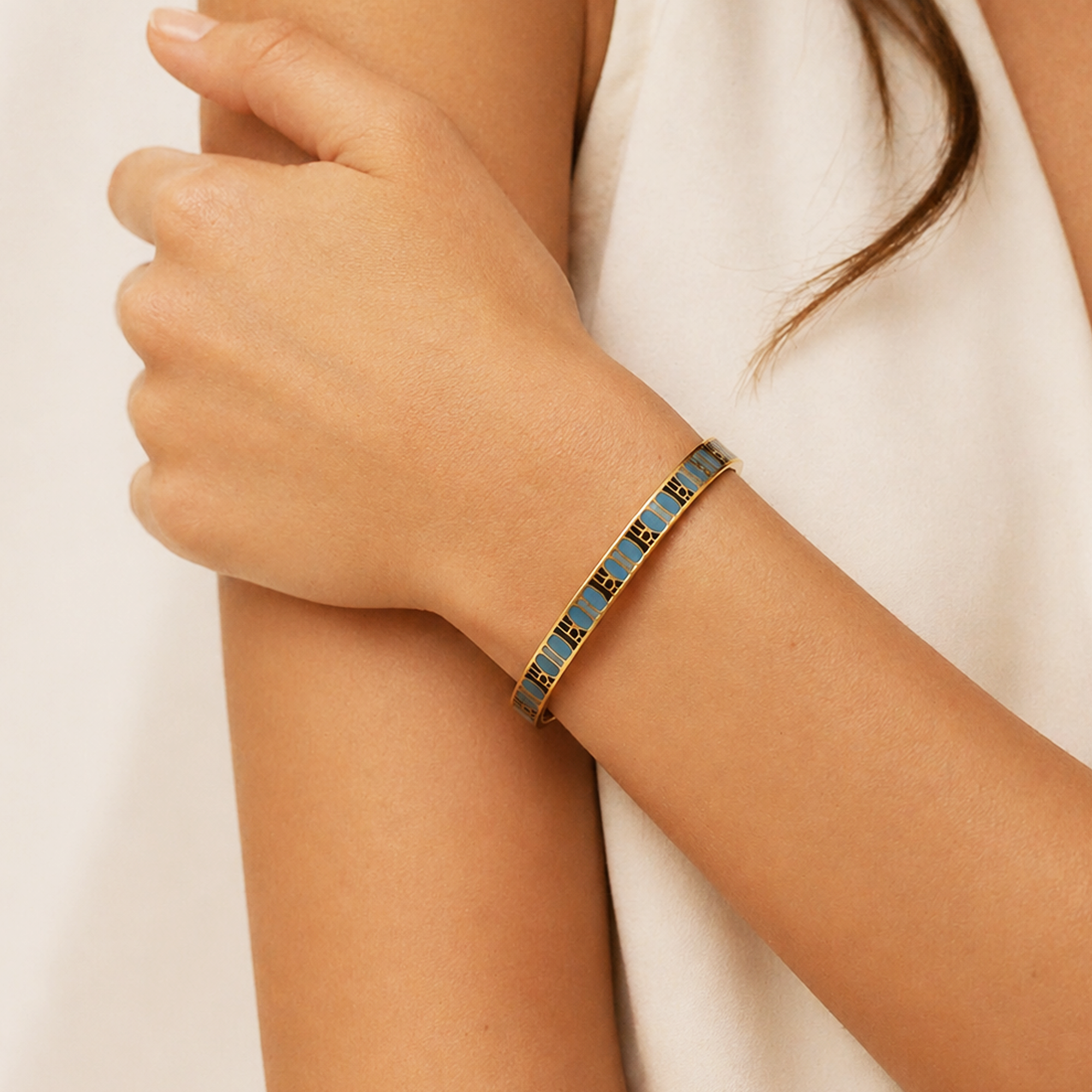 A close-up of a woman's wrist wearing a slim bangle bracelet with a blue and gold geometric pattern, paired with a white sleeveless top.