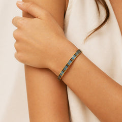 A close-up of a woman's wrist wearing a gold-toned bangle bracelet with blue enamel panels and small gold geometric details, against a soft cream-colored top and neutral background.