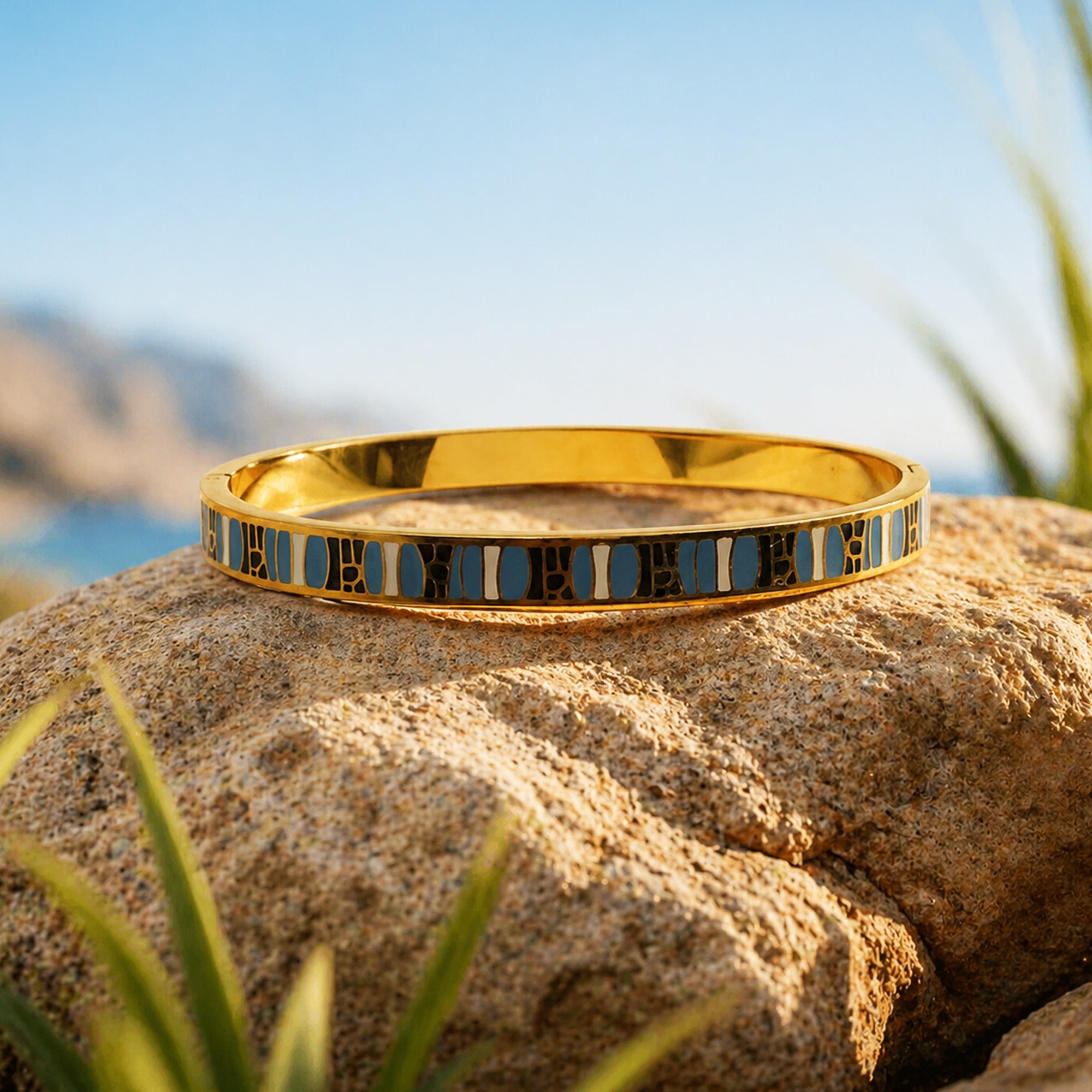 A gold bangle bracelet with a blue and white geometric patterned inlay, resting on a large tan rock with a blurred outdoor background of blue sky and green plants.