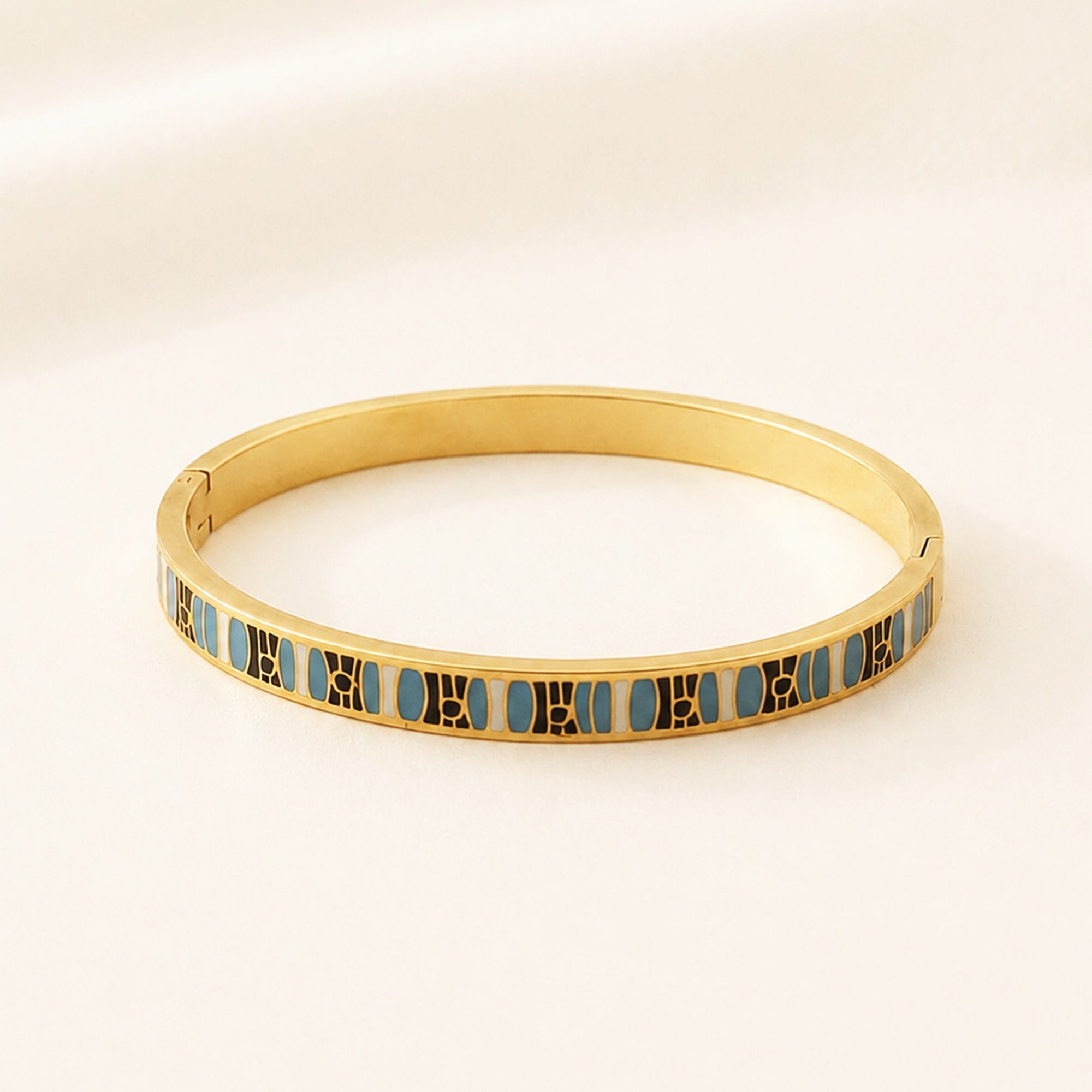 A gold-plated bangle bracelet with a repeating pattern of light blue oval inlays bordered by gold lines, displayed on a clean, light beige background.