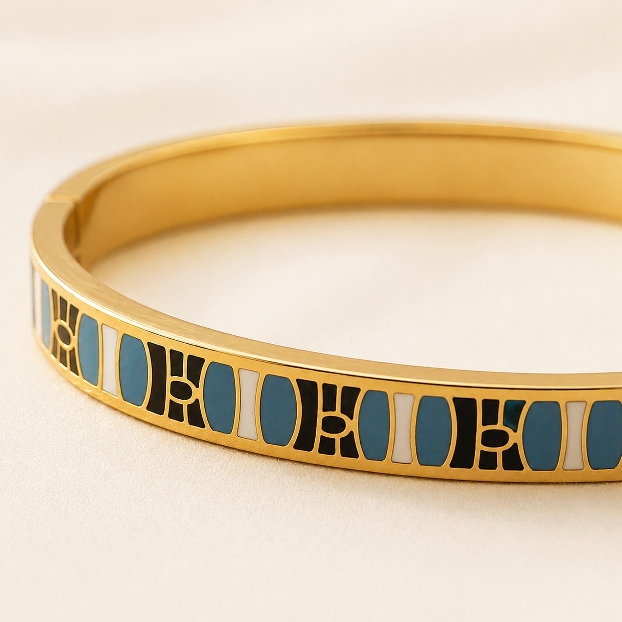A close-up of a gold bangle bracelet featuring a geometric inlay pattern of black, white, and blue enamel.