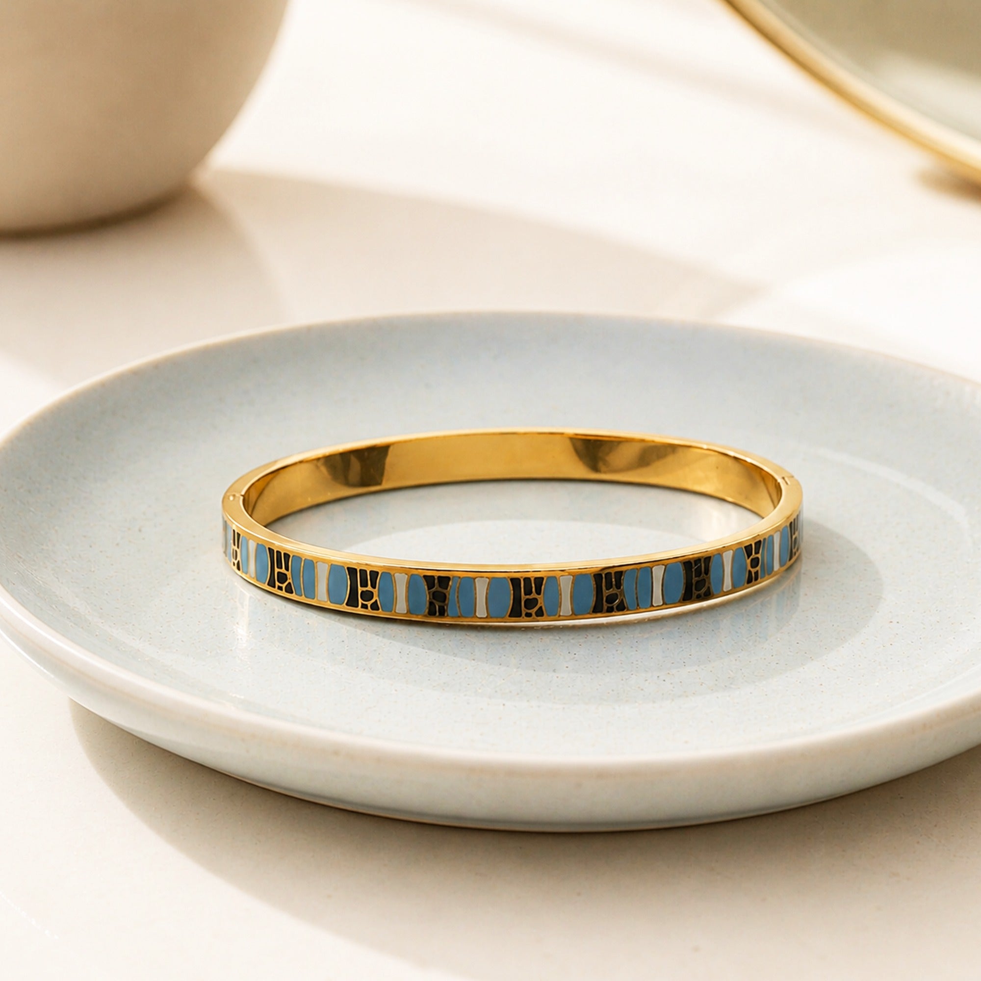 A gold bangle bracelet with a row of light blue and black enamel rectangular details, resting on a light blue ceramic dish.