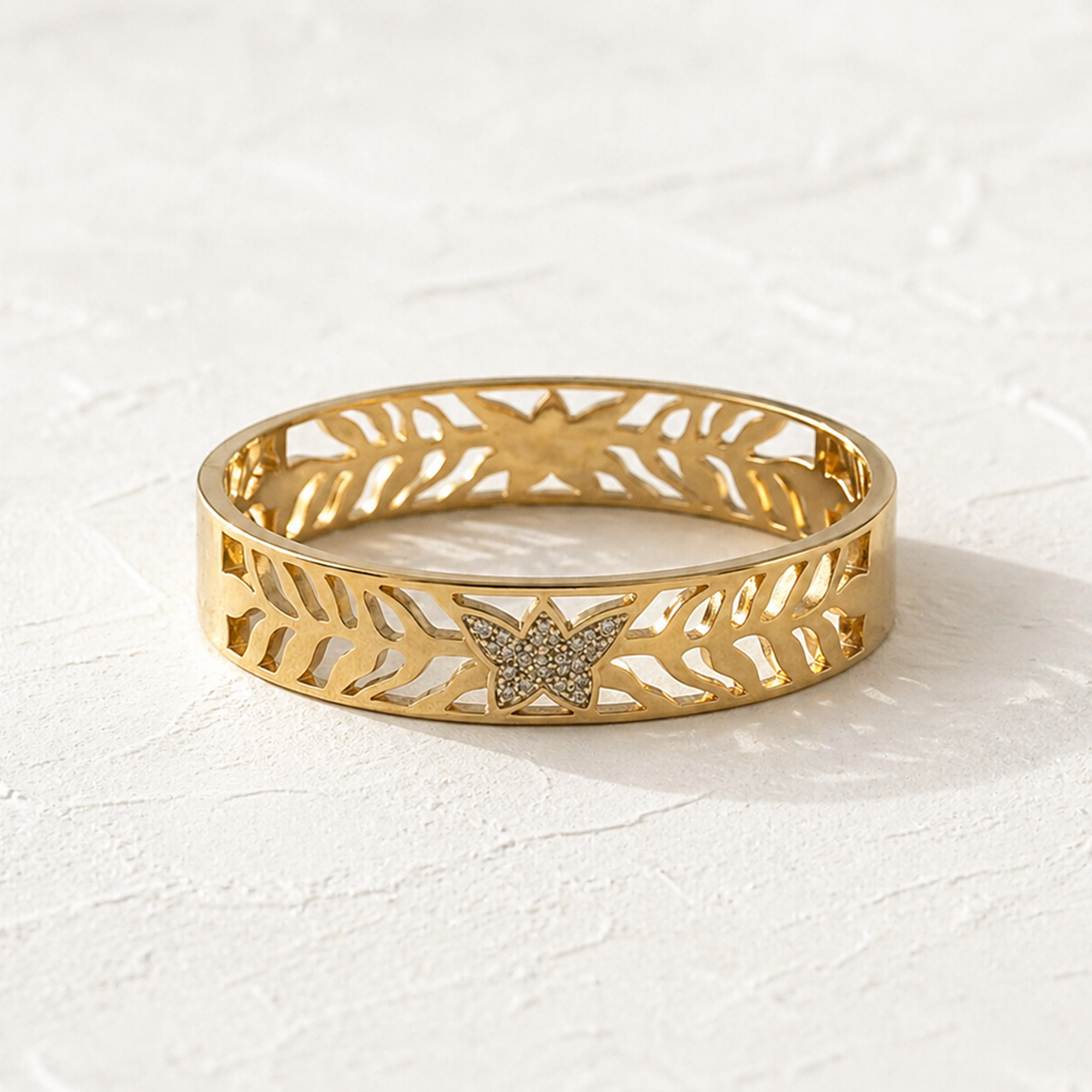 A gold bangle bracelet with a butterfly motif and openwork leaf patterns, set against a textured white background.