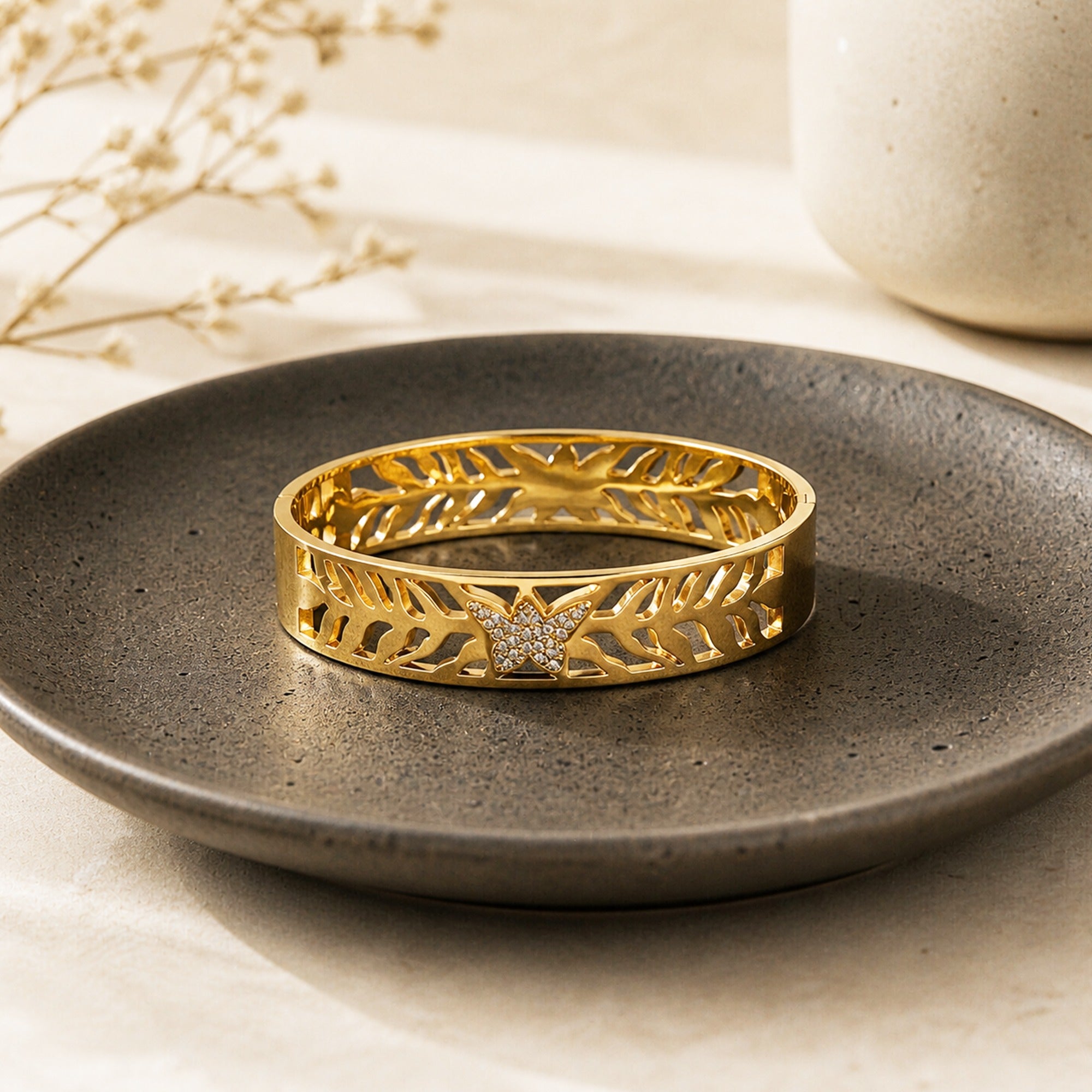 A gold bangle bracelet with an intricate leaf pattern and a central star-shaped crystal cluster, displayed on a dark gray ceramic plate.