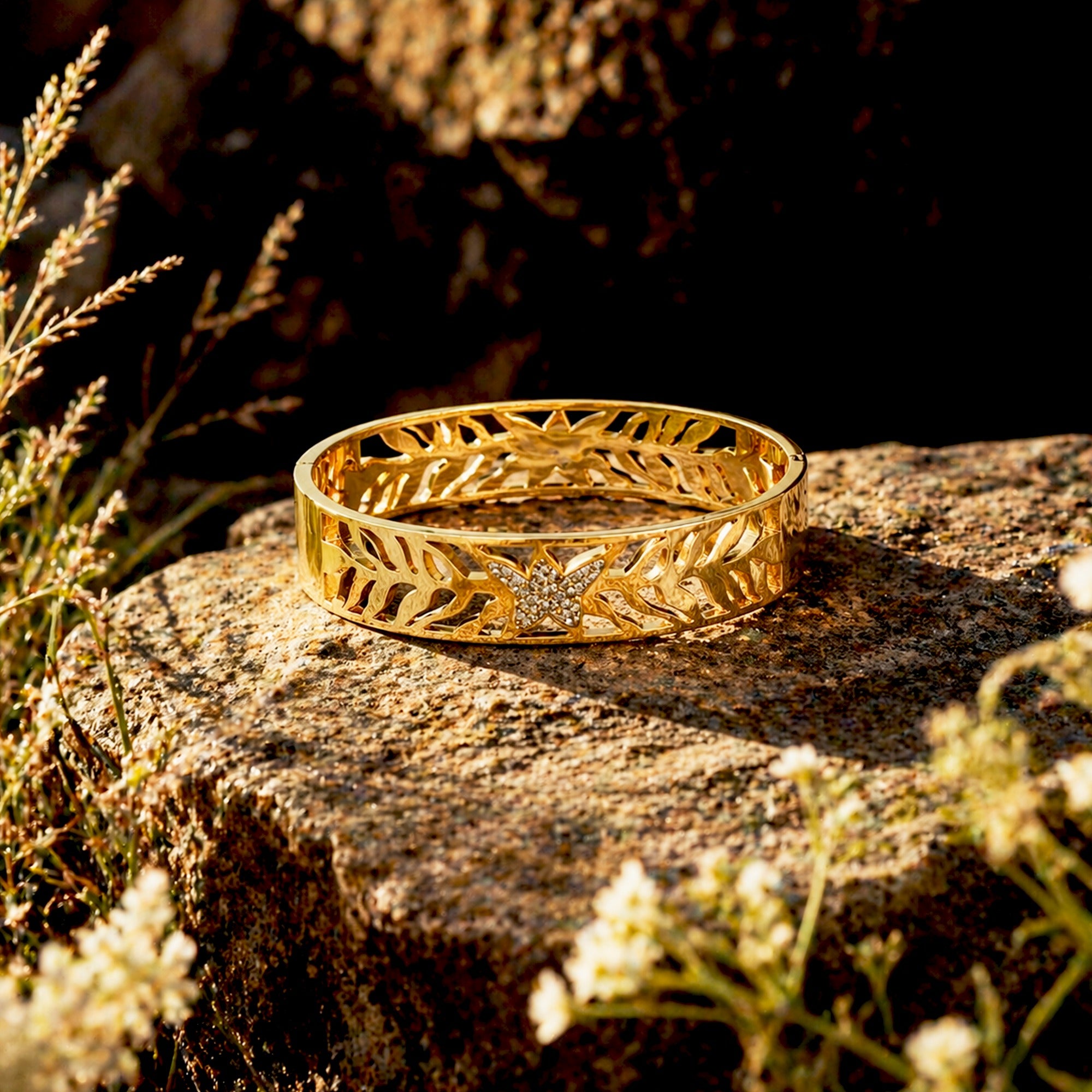 A gold bangle bracelet with an intricate leaf filigree pattern and a central diamond, resting on a textured stone surrounded by wildflowers.