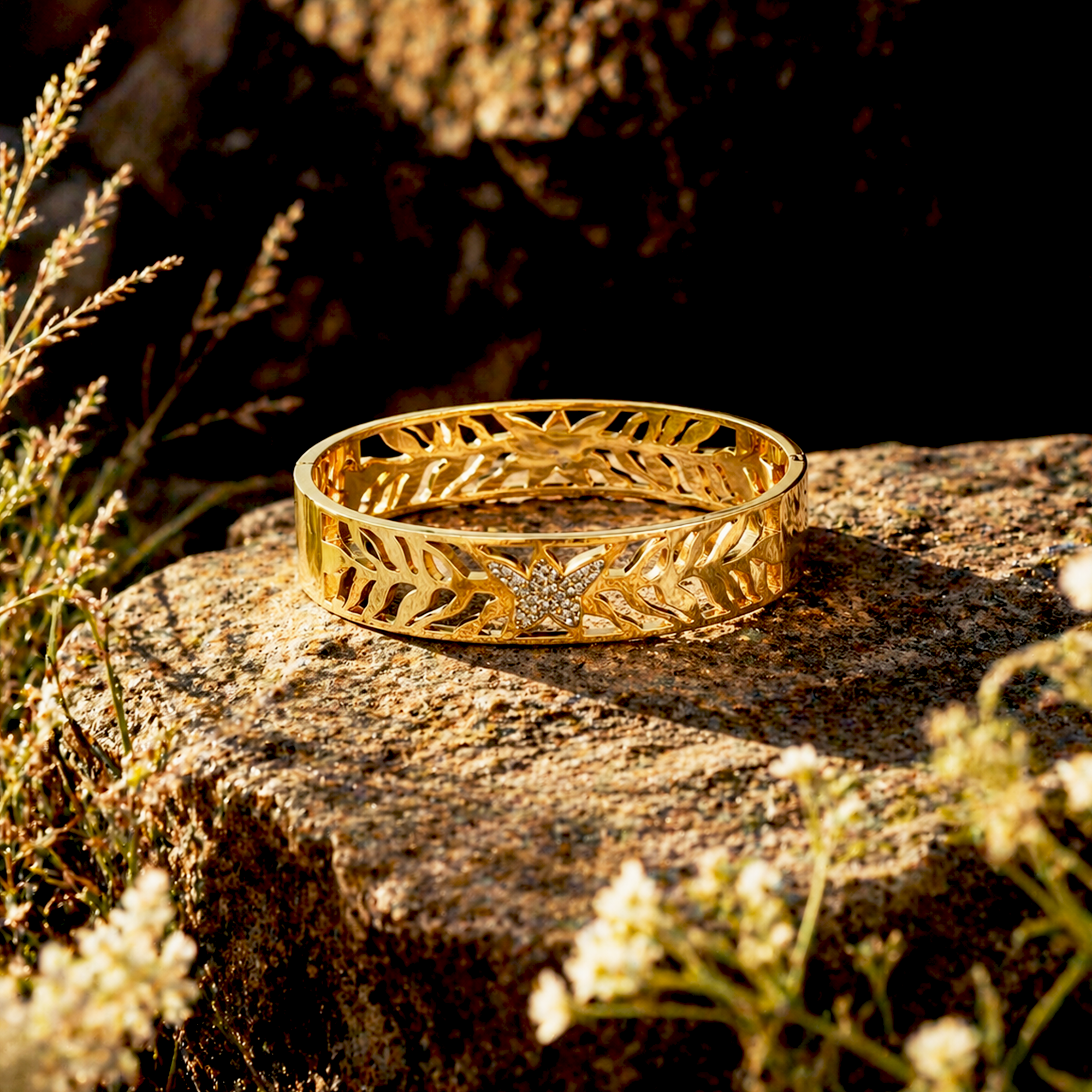 A gold bangle bracelet with an intricate leaf filigree pattern and a central diamond, resting on a textured stone surrounded by wildflowers.