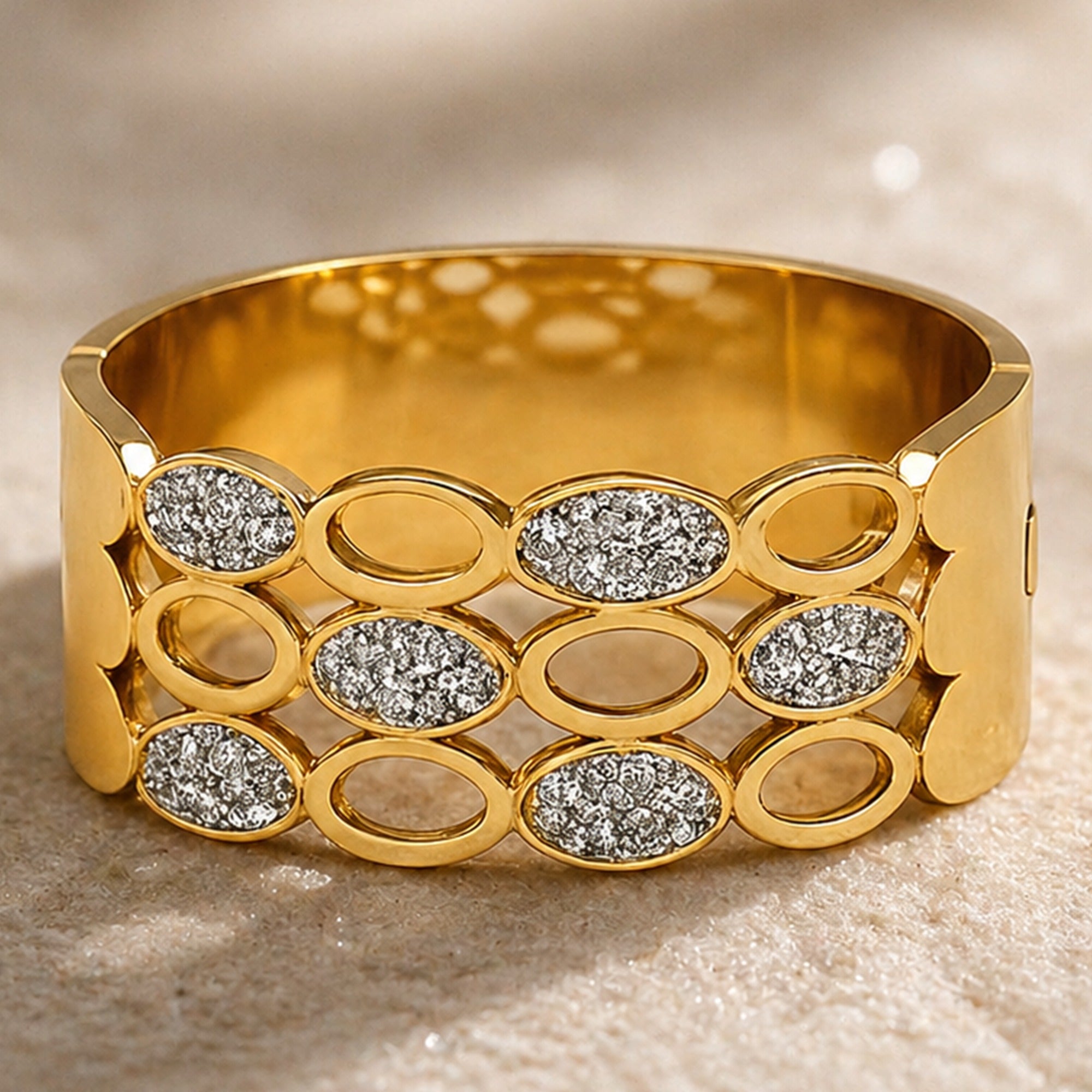 A wide gold bangle bracelet featuring a grid pattern of oval links and diamond-shaped pavé-set stones, photographed on a textured beige surface.