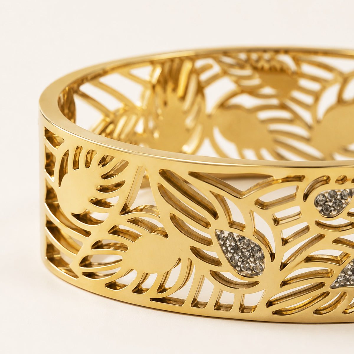 A close-up of a gold-toned cuff bracelet featuring an intricate openwork leaf pattern and small teardrop-shaped crystal accents, set against a clean white background.