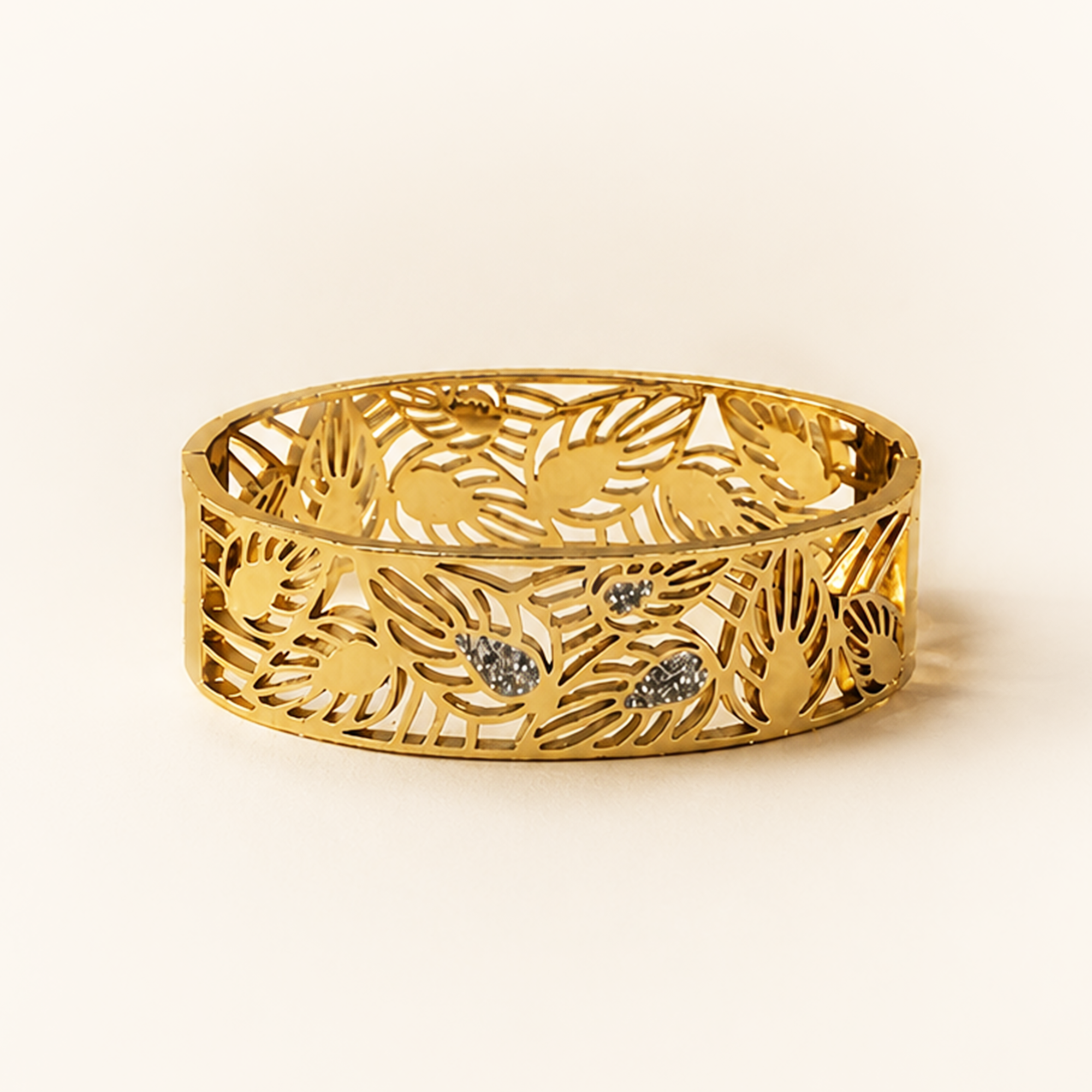 A gold-colored filigree bangle bracelet with an intricate leaf and floral cutout pattern, accented with three small sparkling clear stones, set against a warm cream background.