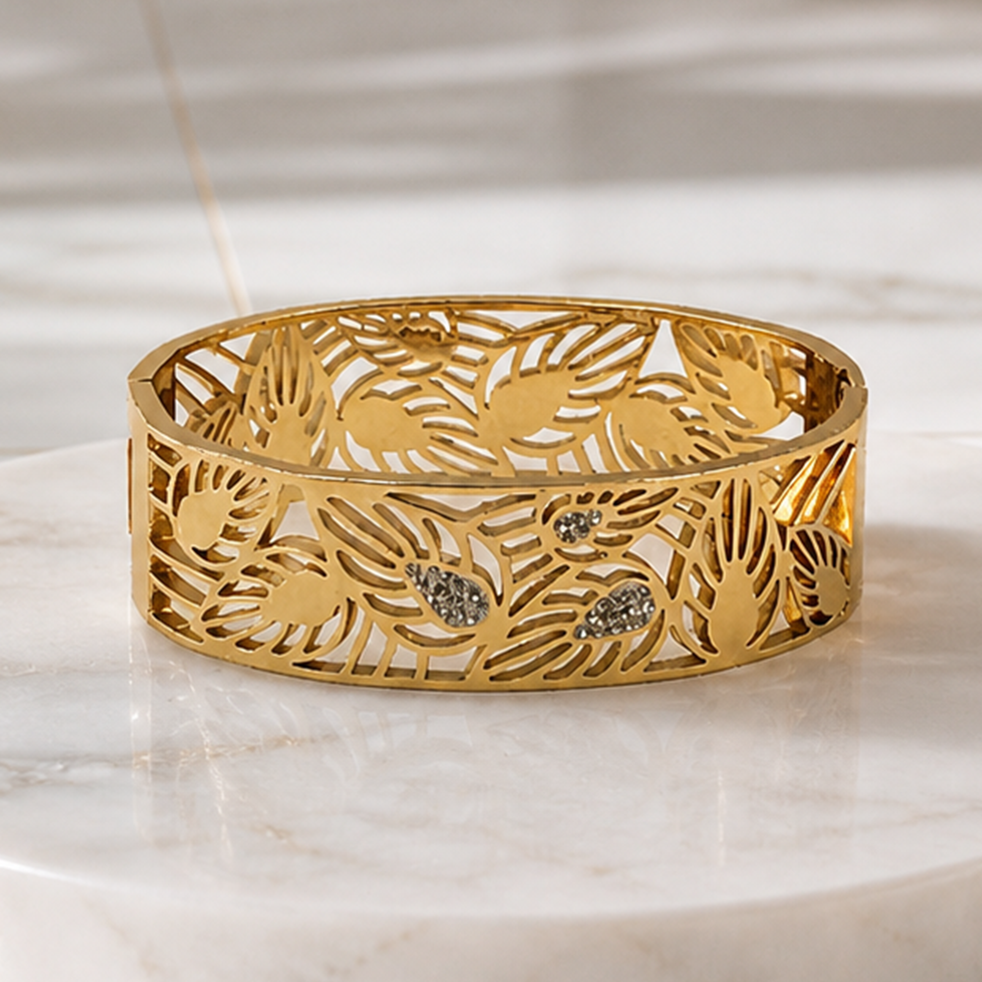 A gold-toned openwork cuff bracelet with an intricate leaf pattern and small clear stone accents, displayed on a light marble surface.