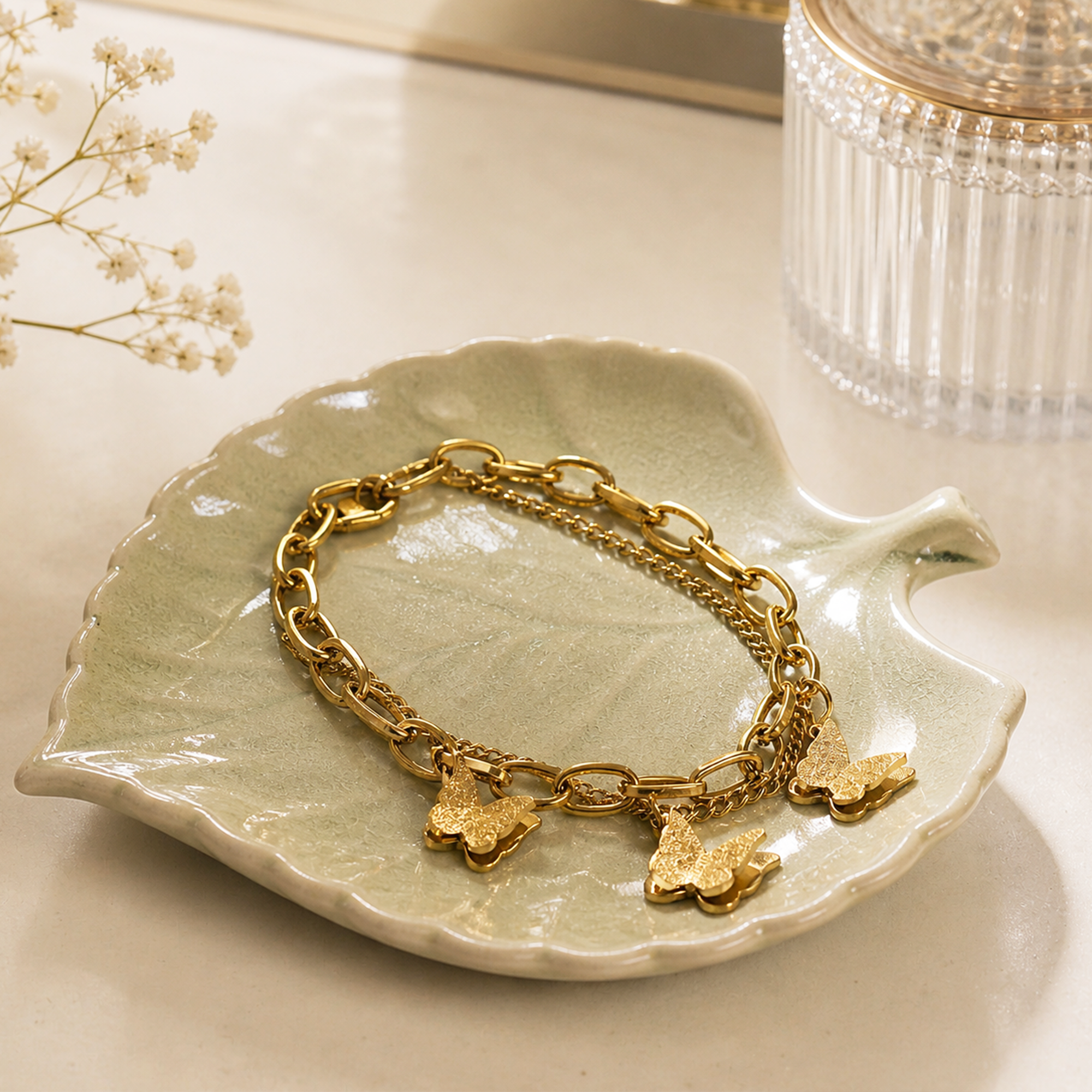 A gold-tone chain bracelet with three textured butterfly charms, displayed on a light green ceramic leaf-shaped dish.