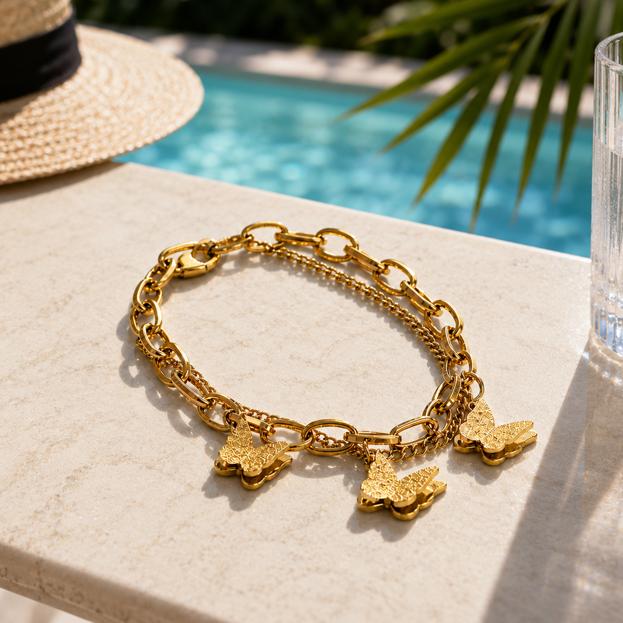 A gold-tone chain bracelet with three textured butterfly charms, displayed on a light beige stone surface with a swimming pool, palm leaves, and a straw hat in the background.