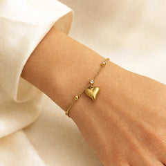 A delicate gold chain bracelet with a small heart charm and tiny bead accents, shown worn on a person's wrist with a white sleeve visible.