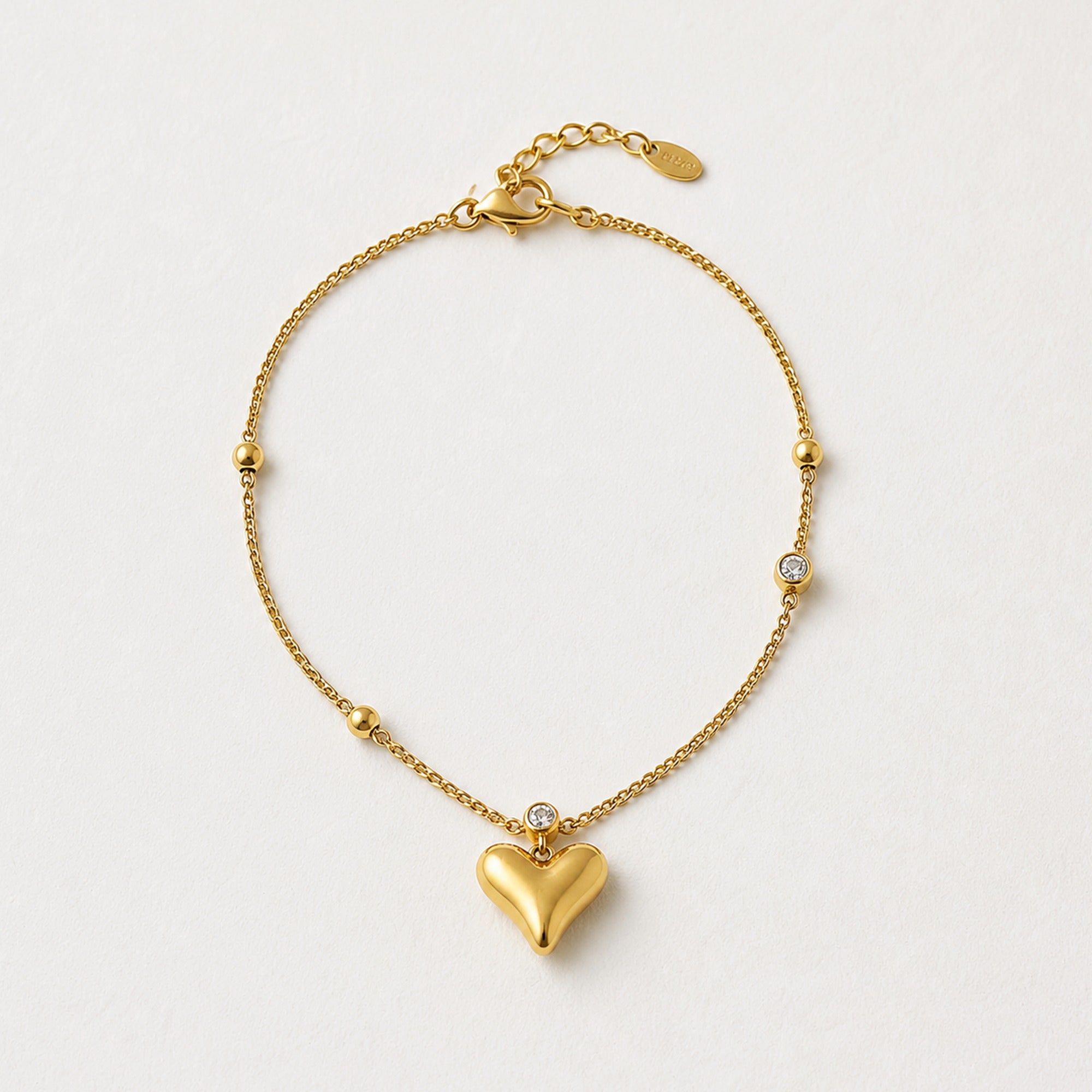 A delicate gold chain bracelet with a polished heart pendant, small gold beads, and a single clear crystal accent, displayed on a clean off-white background.