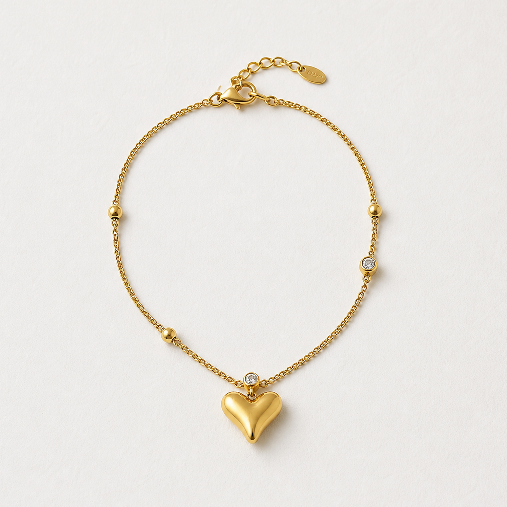 A delicate gold chain bracelet featuring a prominent heart-shaped pendant at the center, small gold beads spaced along the chain, and a tiny clear stone accent, photographed on a clean off-white background.