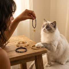 A person holds up a black beaded bracelet with a gold starfish charm next to a fluffy white and gray cat with blue eyes, who is reaching toward bracelets on a small wooden table.