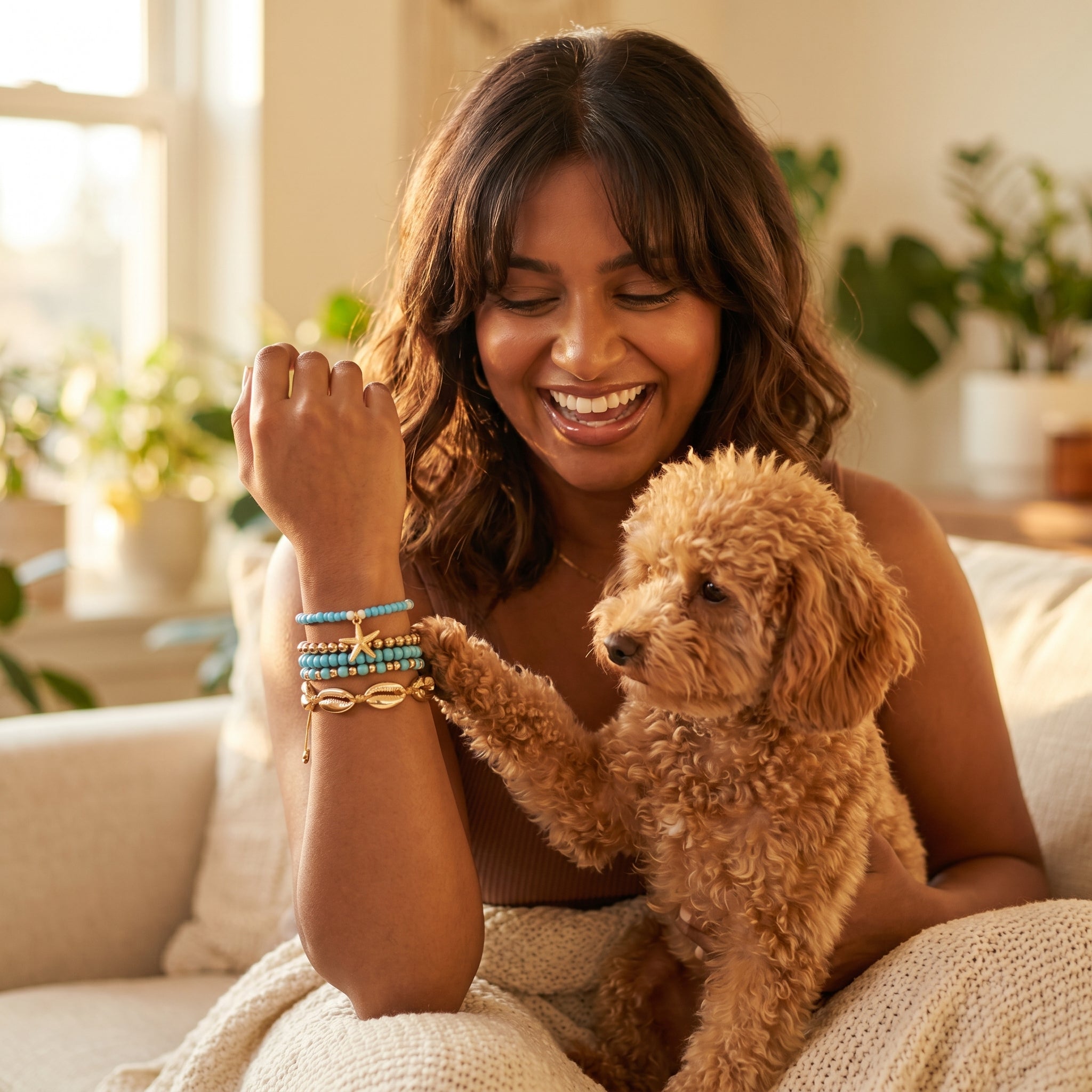 A woman smiling on a couch while wearing colorful beaded bracelets, with a small fluffy brown dog resting on her lap and touching her wrist to show the bracelets. The scene is set in a bright, cozy living room with plants in the background.