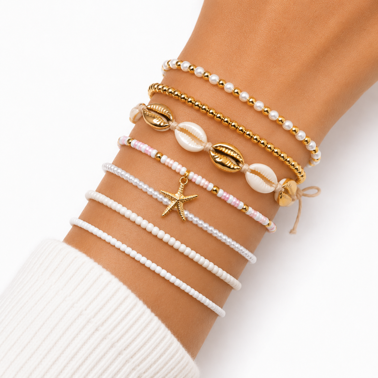 A close-up of a person's wrist wearing six delicate bohemian-style beaded bracelets. The bracelets include white pearl-like beads, gold-tone beads, natural cowrie shells, a pink-and-white beaded strand with a gold starfish charm, and a white beaded bracelet with a small gold bell.