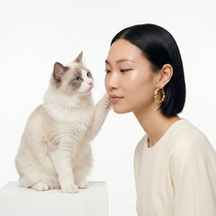 A woman with short black hair wearing gold hoop earrings and a cream top sits beside a fluffy cream and gray cat that is touching her face, against a clean white background.