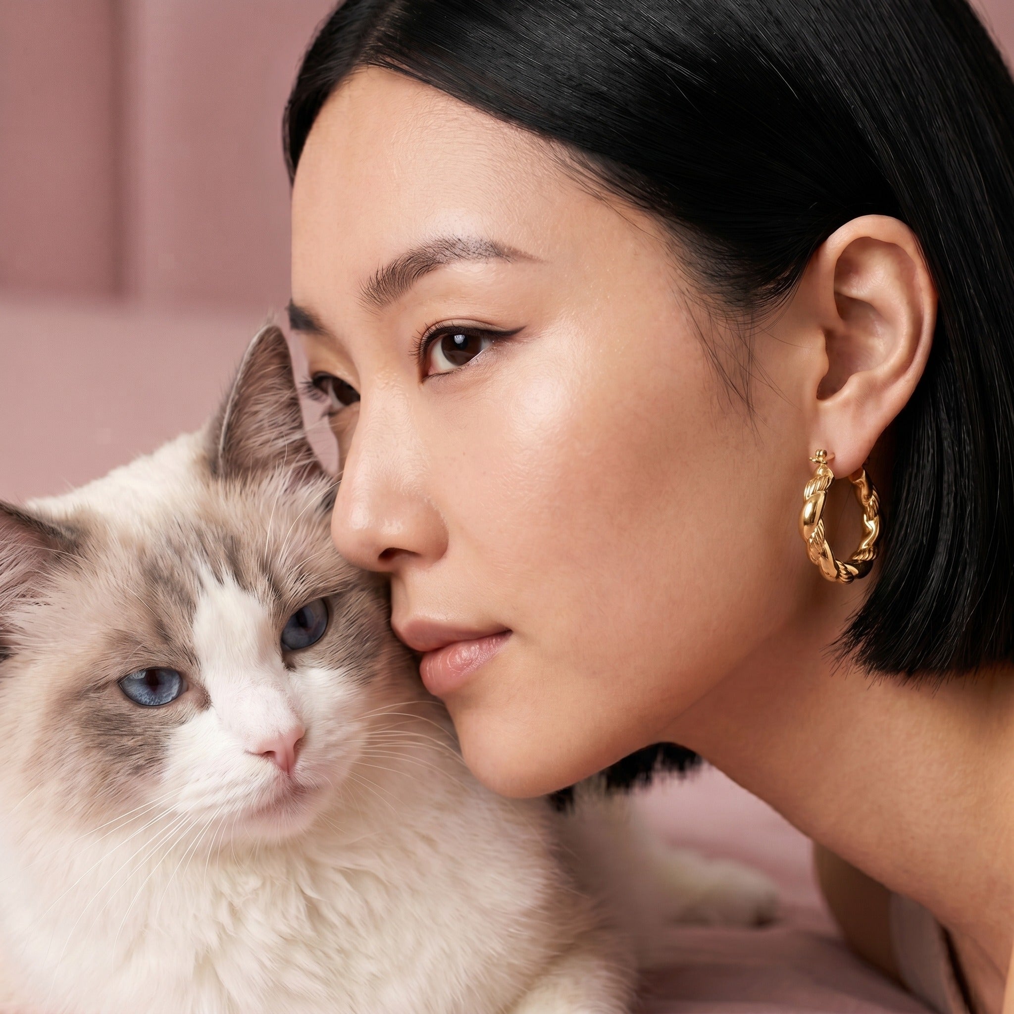 A close-up lifestyle shot of a woman with short black hair wearing a gold twisted hoop earring, resting her head against a fluffy white and gray cat. The background is a soft, solid dusty pink.