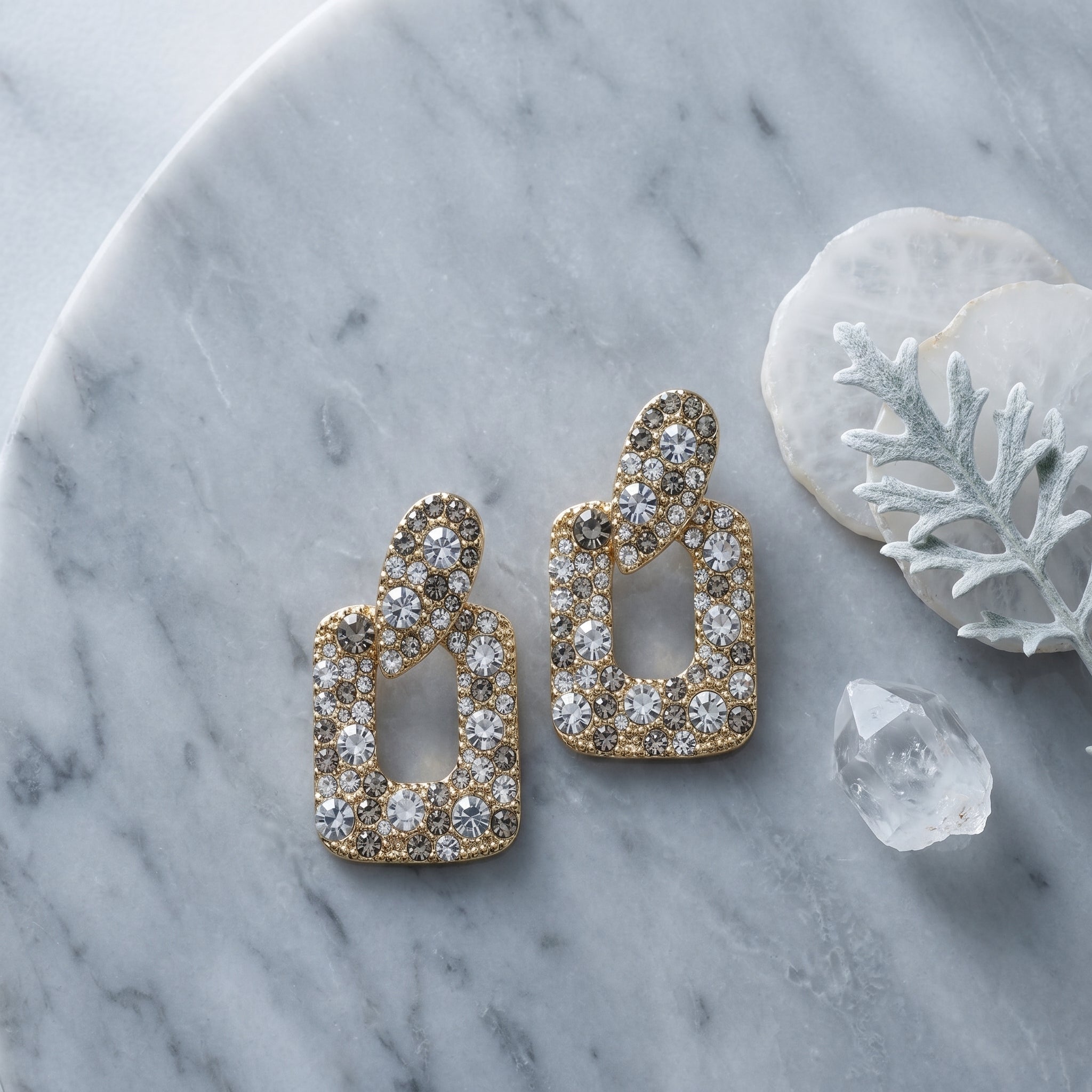 A pair of gold-toned statement earrings adorned with clear crystals, arranged on a gray marble surface alongside decorative white stones and a white branch.