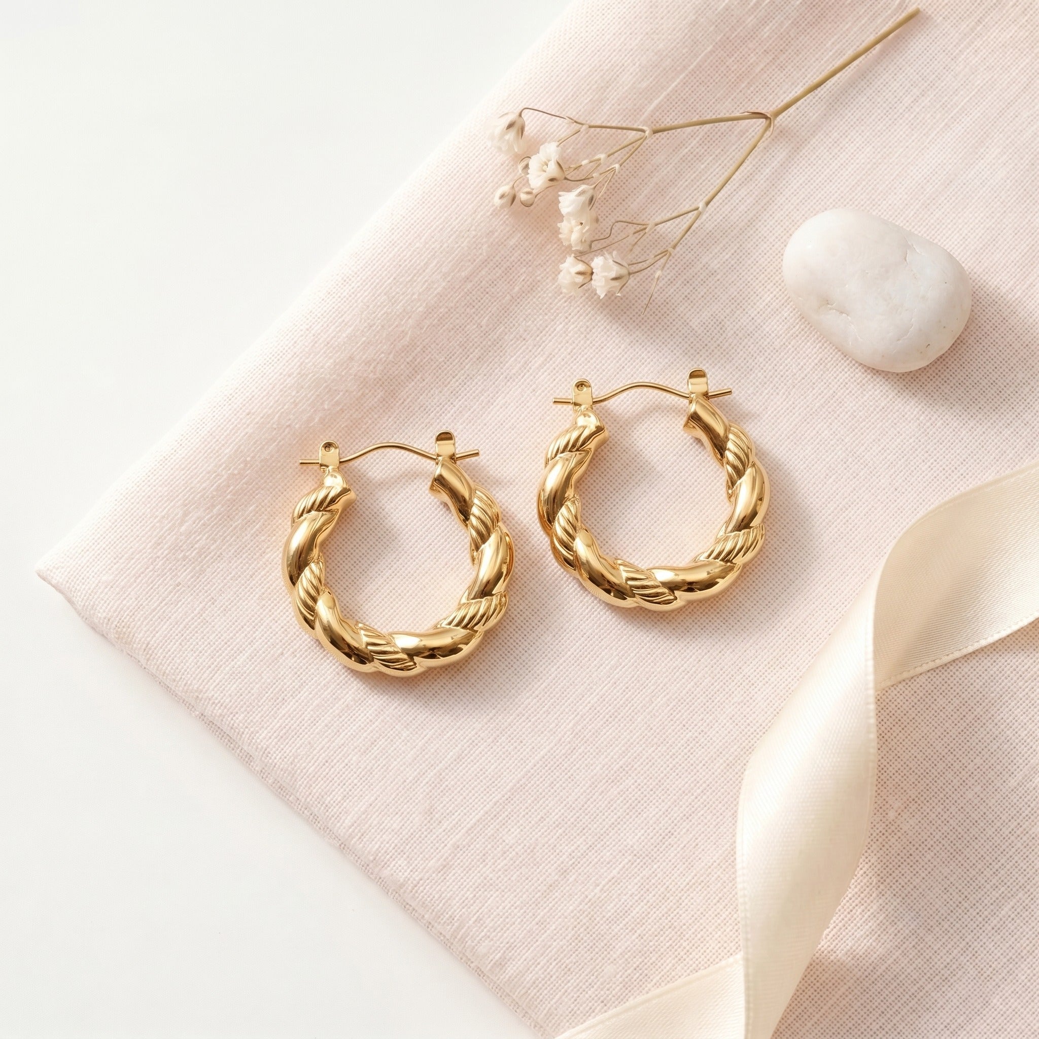 A pair of gold-toned twisted hoop earrings displayed on a pale blush pink fabric surface with dried flowers, a white stone, and a cream ribbon.