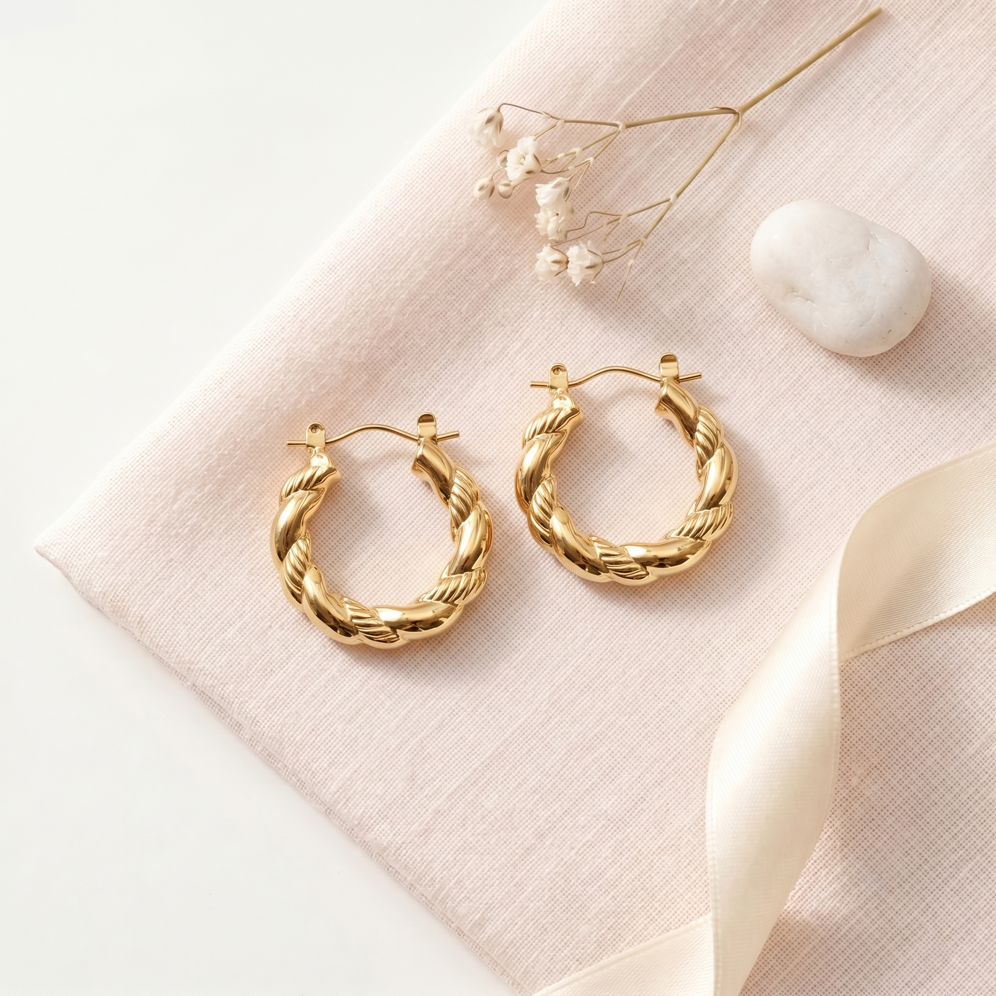 A pair of gold-toned twisted hoop earrings displayed on a pale pink fabric surface with dried flowers, a white stone, and a cream ribbon.
