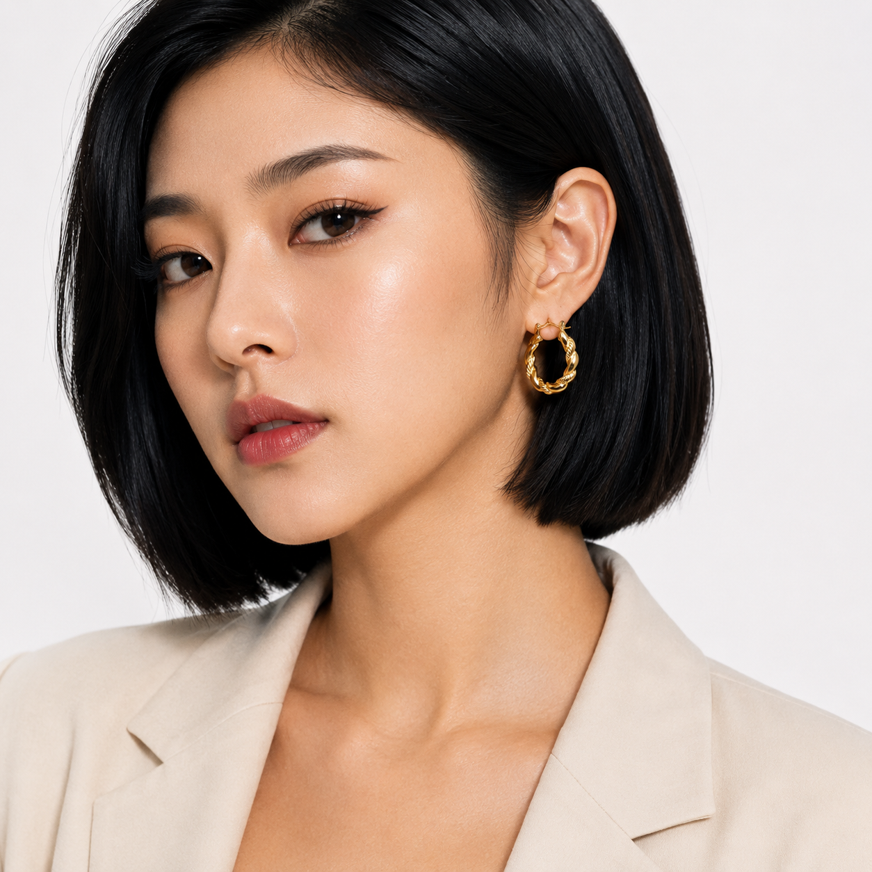 A close-up of a woman with a short black bob hairstyle wearing a textured gold hoop earring, paired with a light beige blazer against a clean white background.