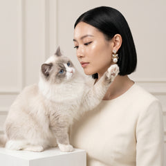 A woman with a short black bob hairstyle and a fluffy cream-colored cat with blue eyes sit together on a white pedestal in a softly lit indoor setting. The woman is wearing a cream-colored top and a pearl bow earring, while the cat reaches toward her face.