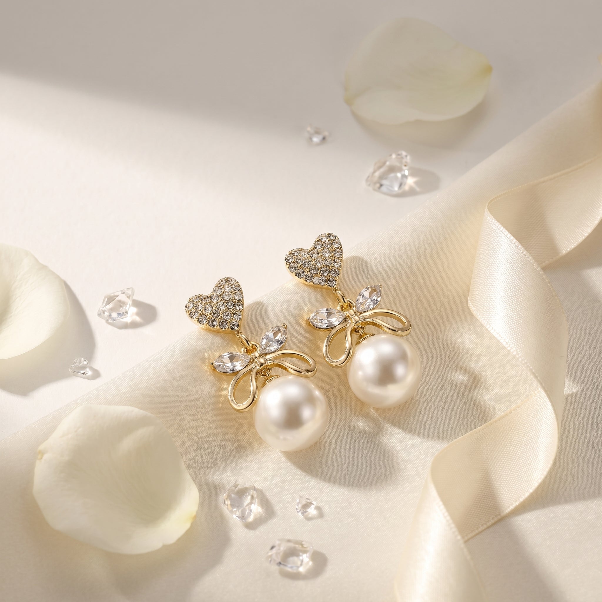 A pair of gold-toned statement earrings with heart-shaped crystal tops, bow-shaped crystal details, and large white pearls, styled on a cream satin fabric with white rose petals and clear crystal chips.