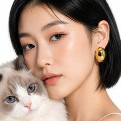 A close-up lifestyle shot of a woman with short black hair wearing a shiny gold oval earring, holding a fluffy gray and white cat against a clean white background.