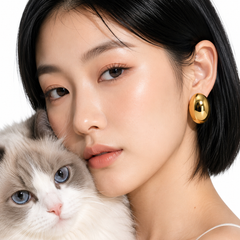 A close-up lifestyle shot of a woman with short black hair wearing a shiny gold oval earring, holding a fluffy gray and white cat against a clean white background.