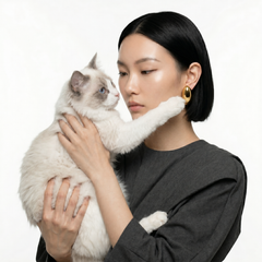 A woman with short black hair holds a fluffy white cat with blue eyes against a plain white background. She is wearing a dark gray long-sleeve top and a gold hoop earring.