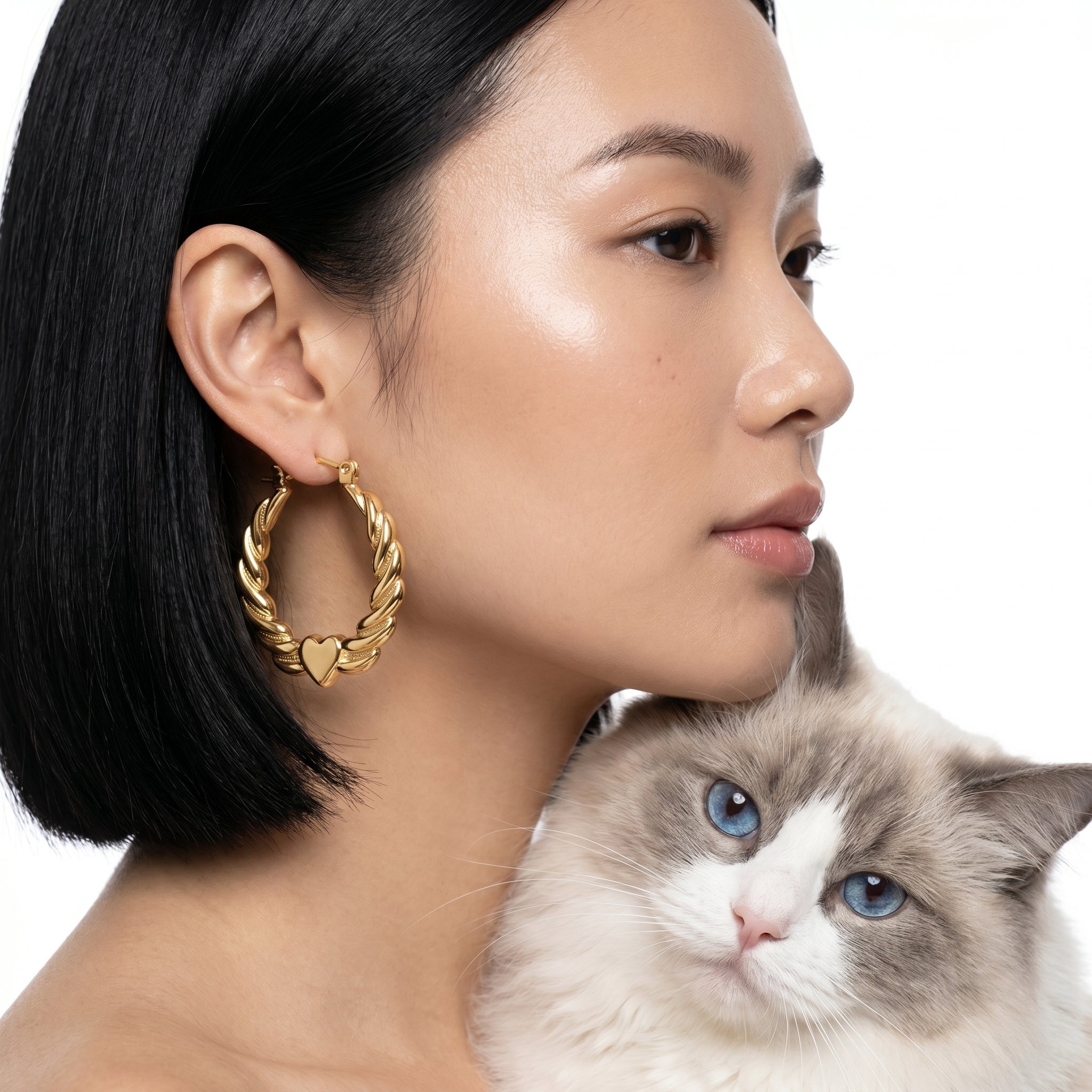 A close-up lifestyle shot of a woman with black hair wearing a gold hoop earring with a small heart detail, with a fluffy white and gray cat resting against her shoulder, set against a solid white background.