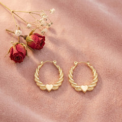 A pair of gold hoop earrings with twisted wing-like edges and a small heart at the center, displayed on a soft pink fabric with dried red roses as props.