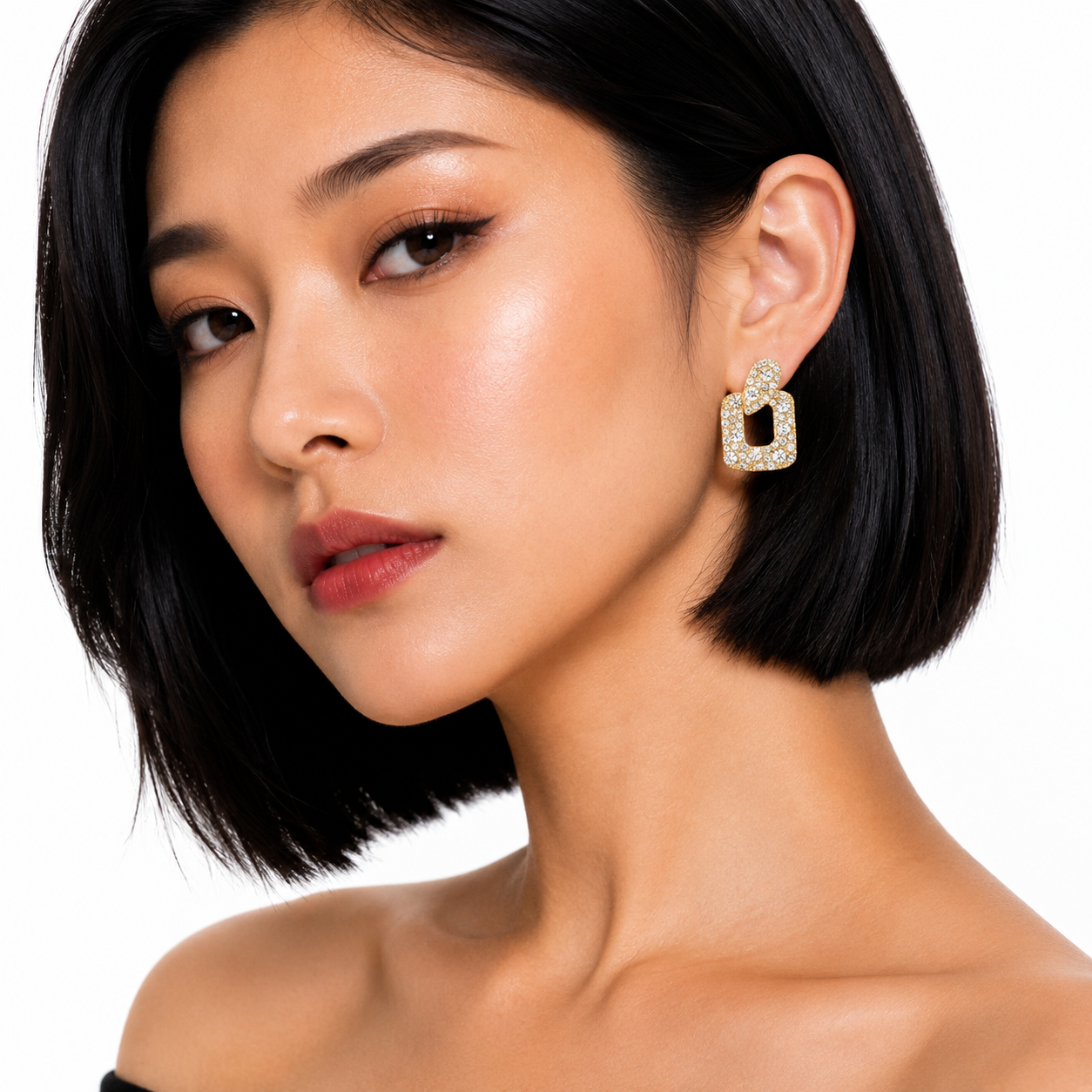 A close-up of a woman with a short black bob hairstyle wearing a gold geometric drop earring adorned with clear crystals, set against a plain white background.