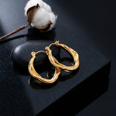 A pair of thick, twisted gold hoop earrings displayed on a polished black stone block with a cotton boll prop, set against a dark reflective background.