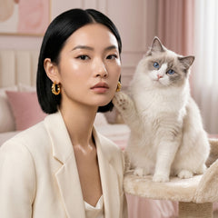 A woman with short black hair and gold hoop earrings wears a cream blazer, sitting beside a fluffy white and gray cat on a cream cat tree in a softly lit pink bedroom.