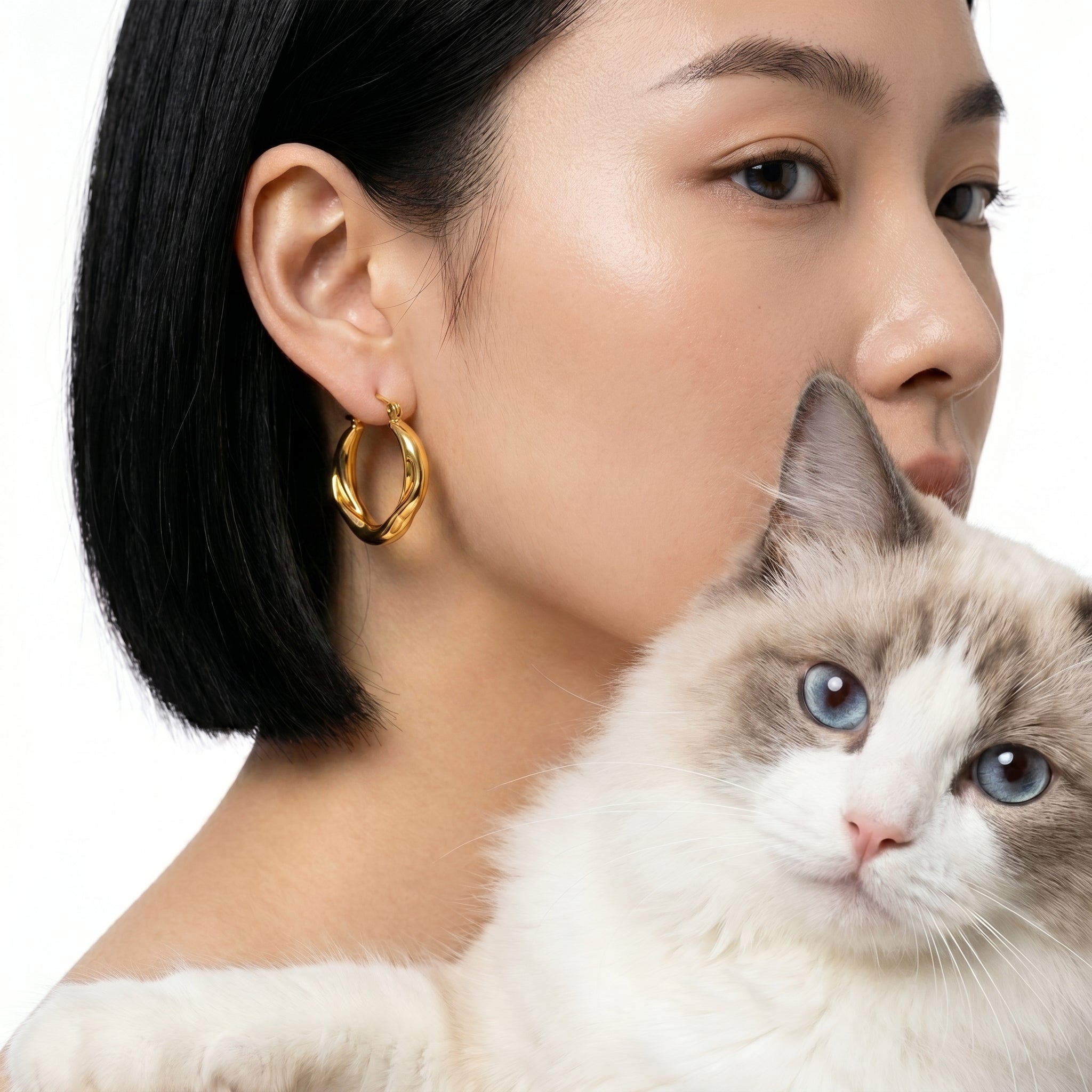 A close-up lifestyle shot of a woman wearing a gold hoop earring, with a fluffy white and gray cat in the foreground against a clean white background.
