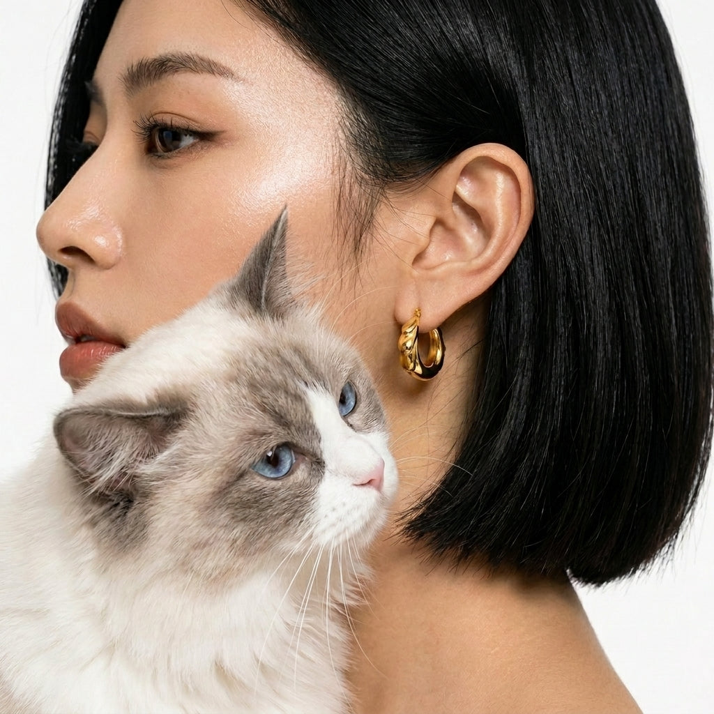 A close-up lifestyle shot of a woman with short black hair wearing a gold hoop earring, with a fluffy white and gray cat resting against her shoulder, set against a clean white background.