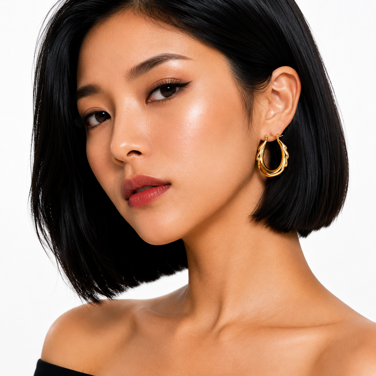 A close-up portrait of a woman with a short black bob hairstyle wearing a textured gold hoop earring, set against a clean white background.