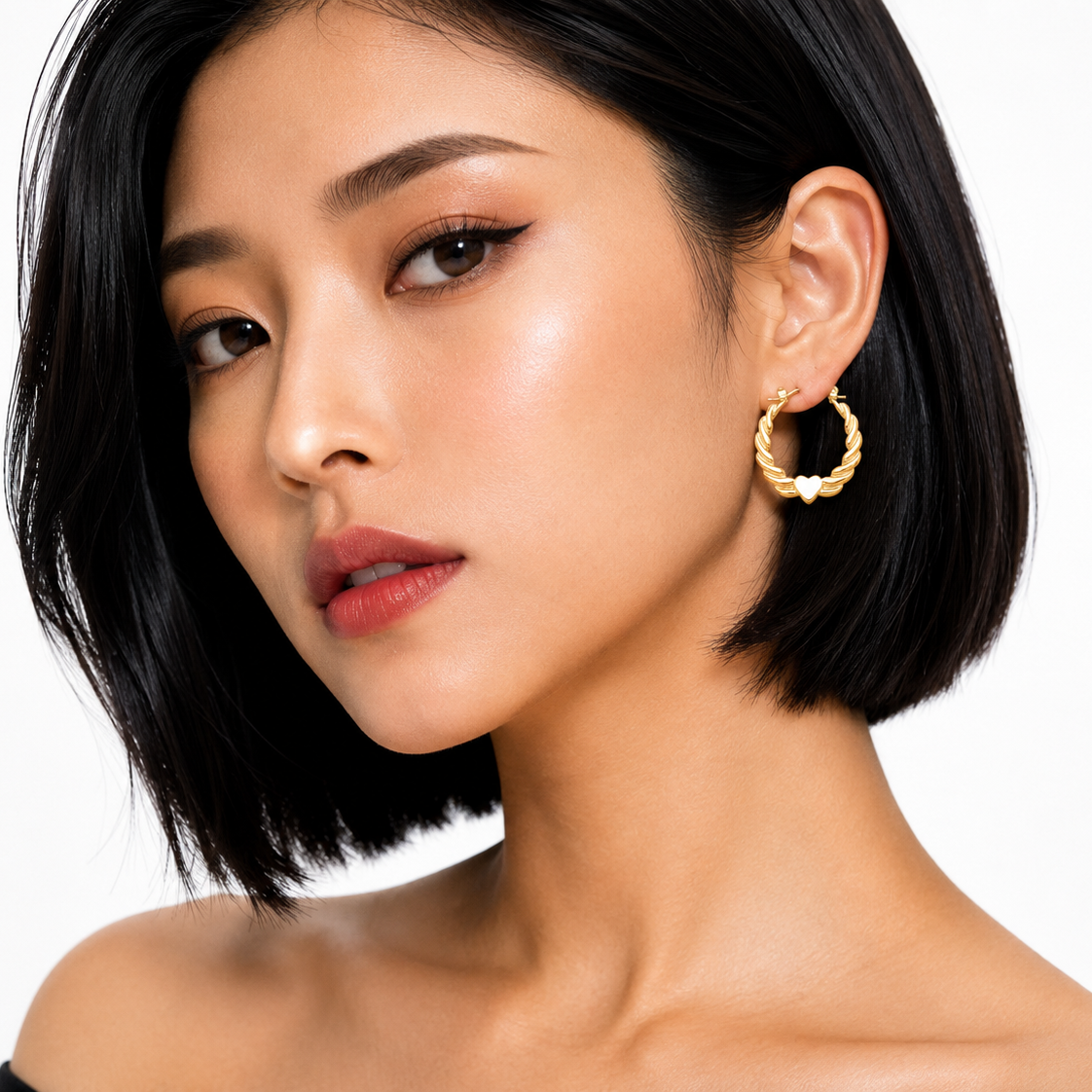 A close-up of a woman with short black hair wearing a textured gold hoop earring with a small white flower detail, set against a clean white background.