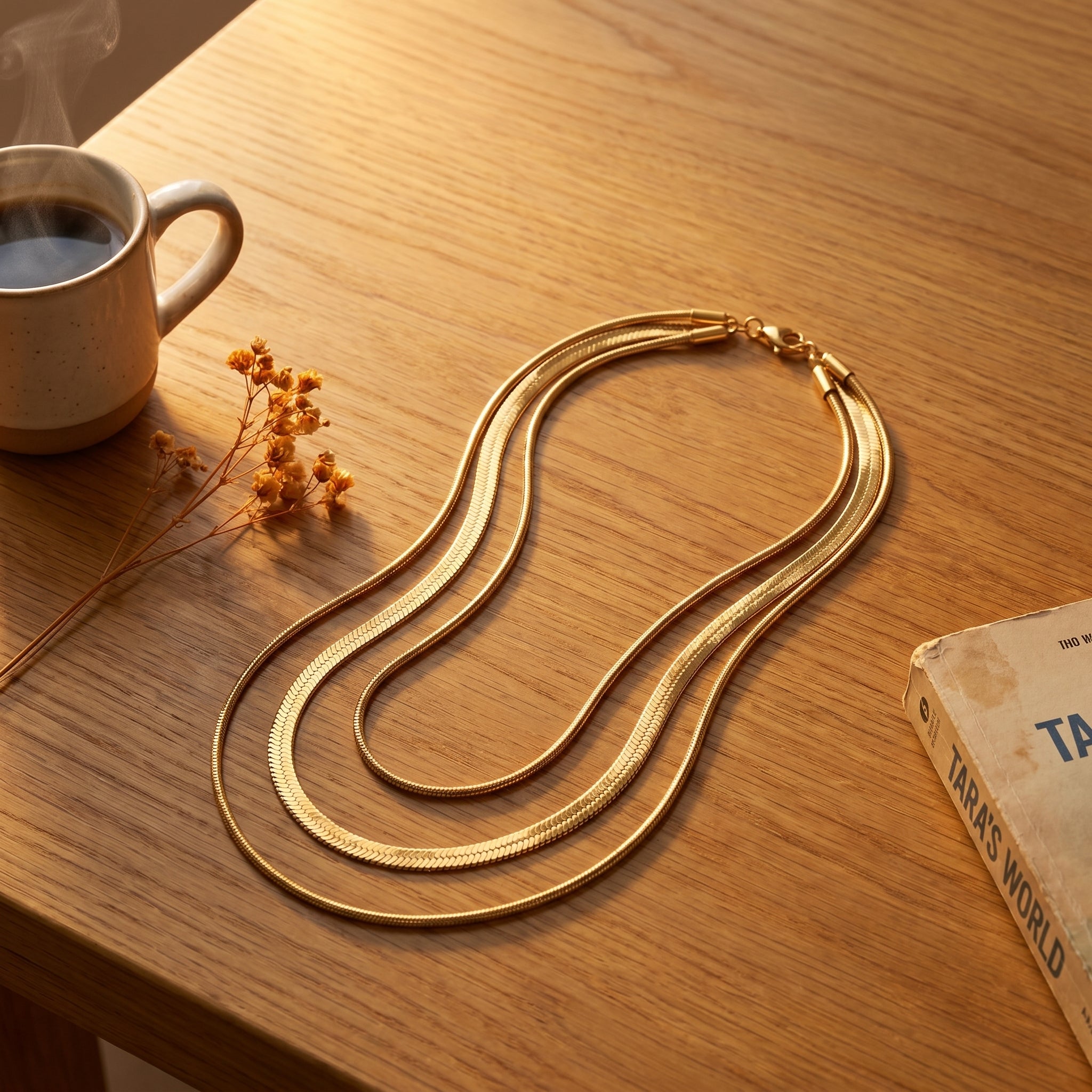 A gold-toned multi-strand snake chain necklace is draped on a warm wooden tabletop, styled with a cup of coffee, dried flowers, and a book.