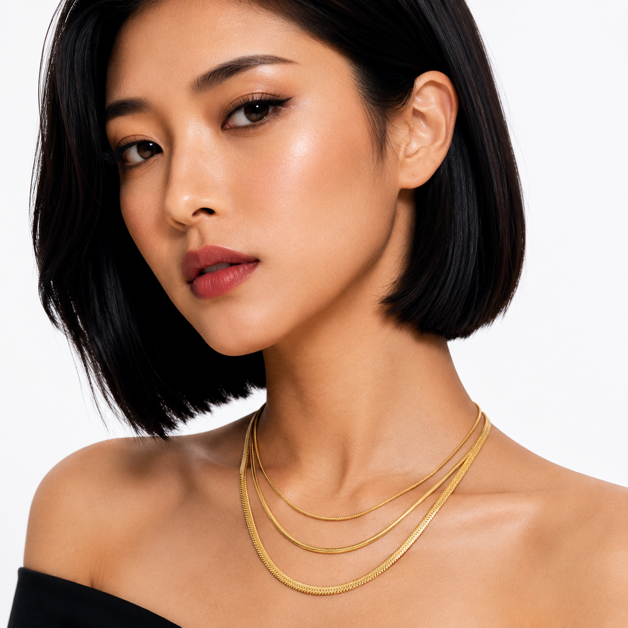 A close-up of a woman with a short black bob hairstyle wearing a delicate layered gold chain necklace against a clean white background.