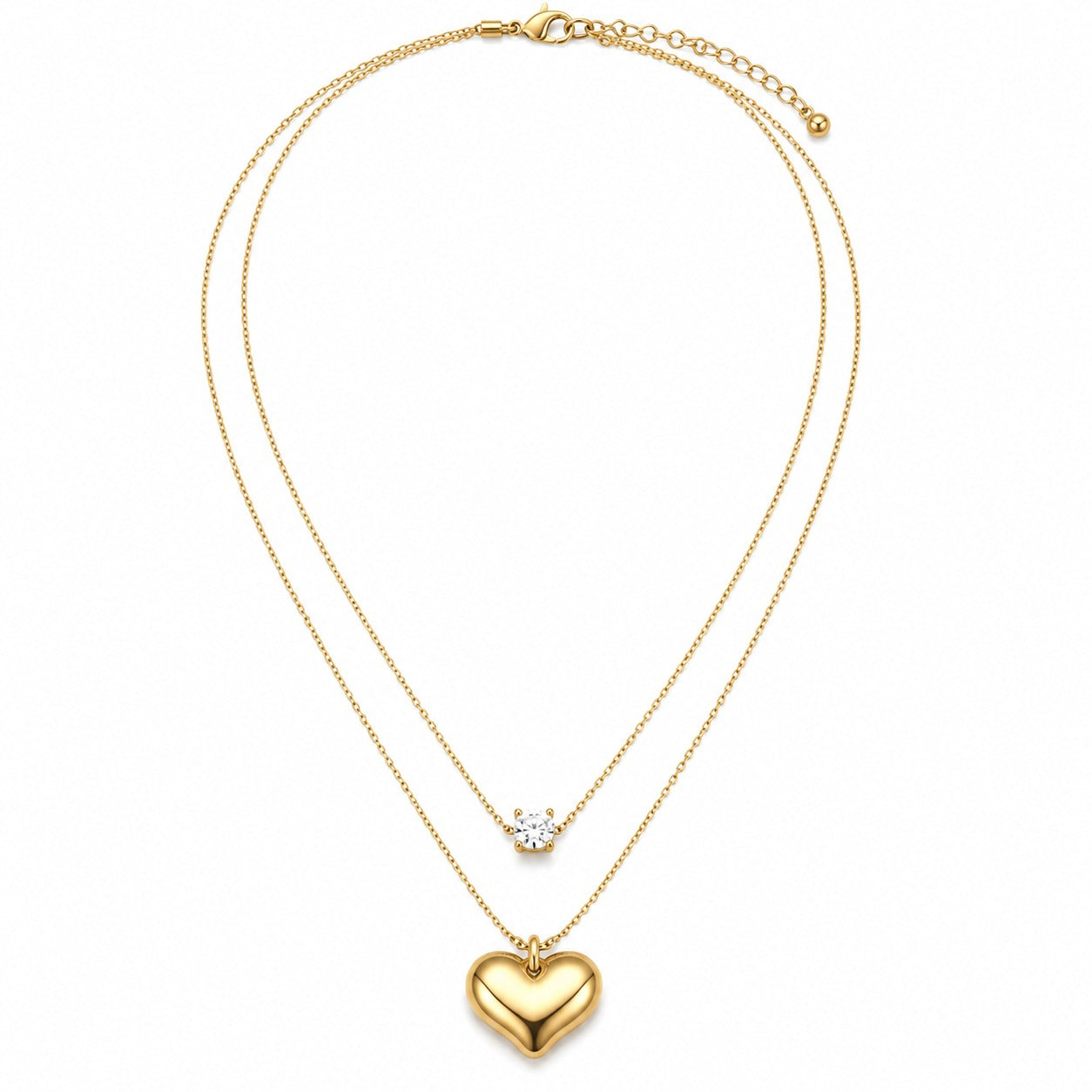 A gold double-layered necklace featuring a polished heart pendant on the longer chain and a small clear gemstone on the shorter chain, displayed on a clean white background.