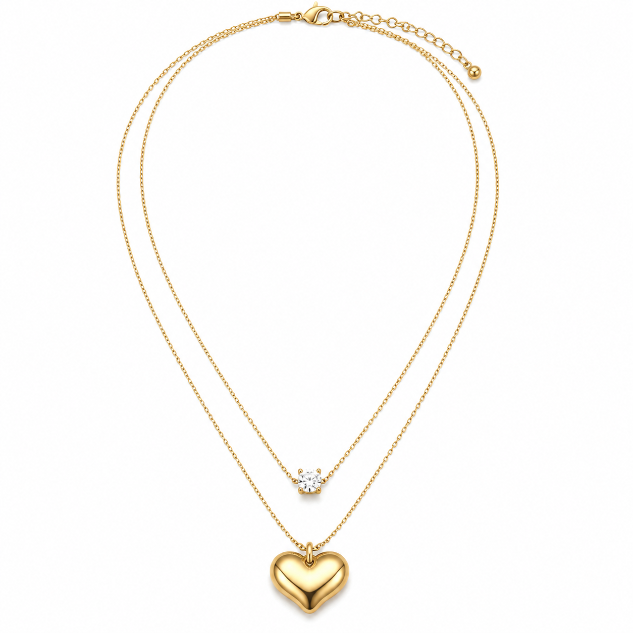 A double-strand gold necklace featuring a polished heart pendant on the longer chain and a small clear gemstone on the shorter chain, displayed against a solid white background.