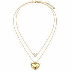 A gold double-layered necklace featuring a polished heart pendant on the longer chain and a small clear gemstone on the shorter chain, displayed on a clean white background.