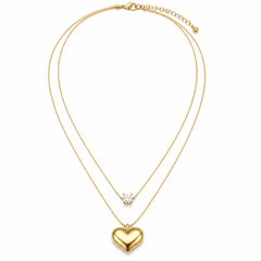 A double-strand gold necklace featuring a polished heart pendant on the longer chain and a small clear gemstone on the shorter chain, displayed against a solid white background.