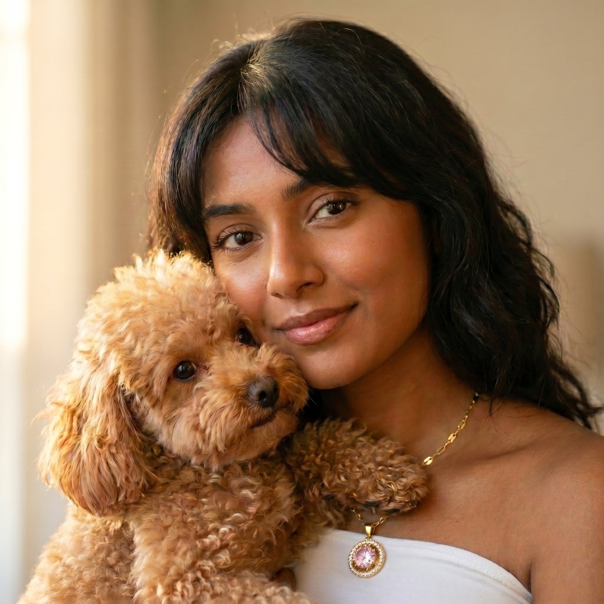 A woman with dark hair smiling while holding a small curly-haired brown dog, wearing a white strapless top and a gold necklace with a pink and white pendant.