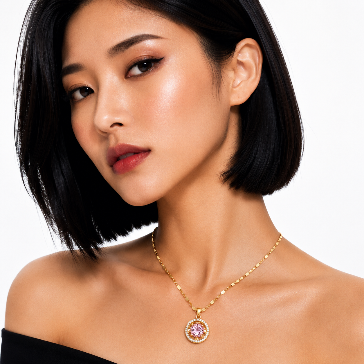 A close-up of a woman with short black hair wearing a delicate gold chain necklace with a round pendant featuring a pink gemstone surrounded by small clear stones, set against a plain white background.
